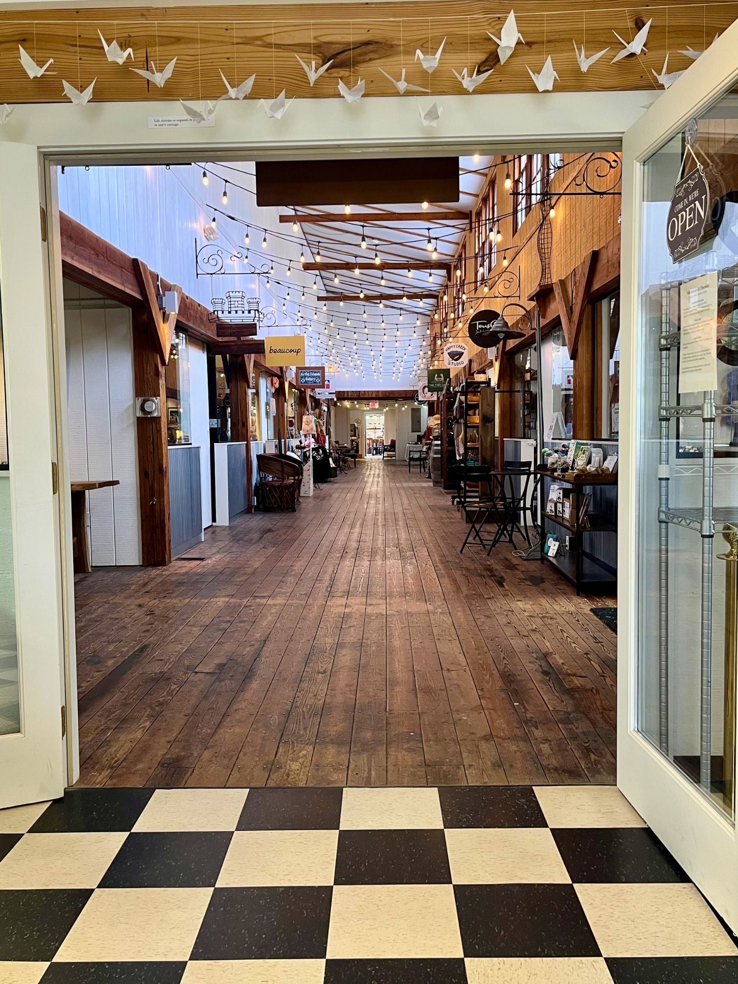 Long hallway with a wooden floor, lined with different stores on both sides. Looking out from Wholehearted Chocolate into hallway. 