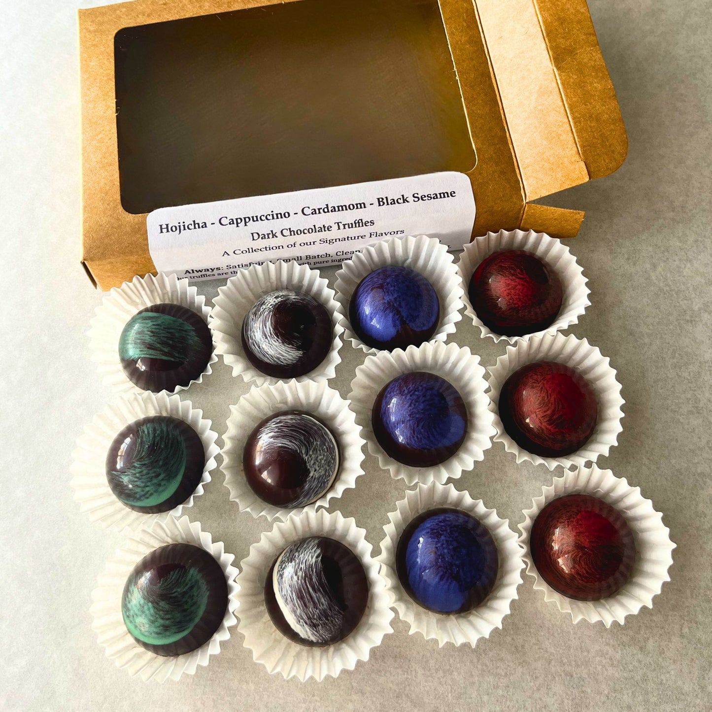 Assorted dark chocolate bonbons in white paper cups near a kraft paper box on a gray surface.