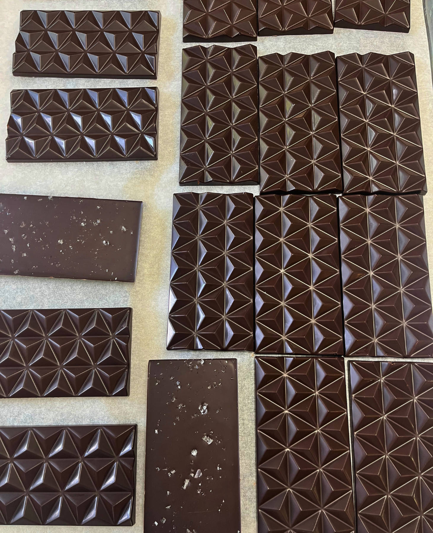 Dark chocolate bars with geometric patterns on a light background, some are topped with flaky sea salt
